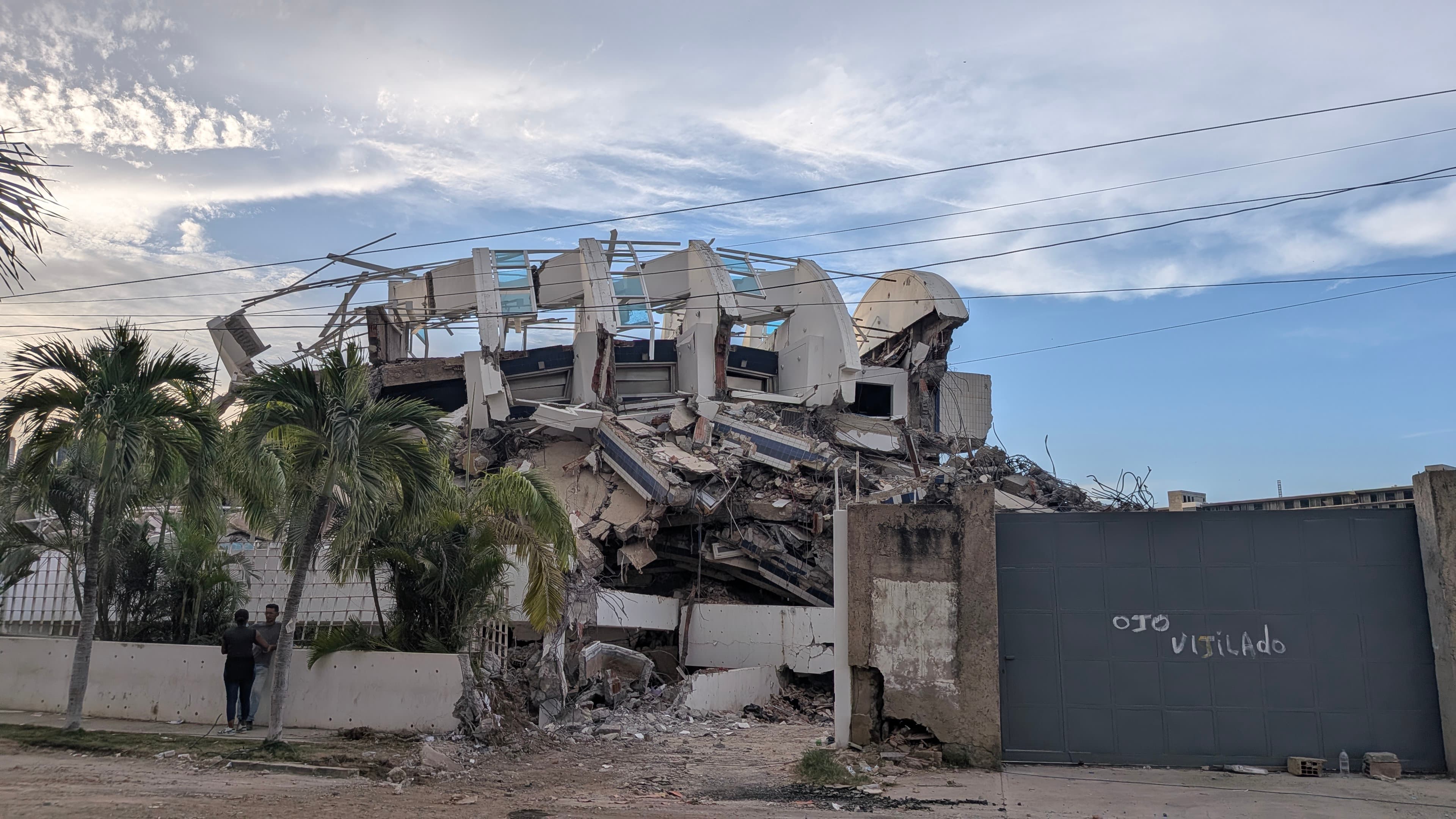 Vecinos observando desde la calle un edificio con el techo colapsado, junto a un portón con la inscripción 'ojo vigilado'.